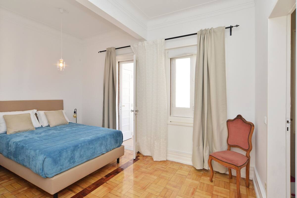 A bedroom with a bed, a chair, a window with curtains, and a hanging light fixture.