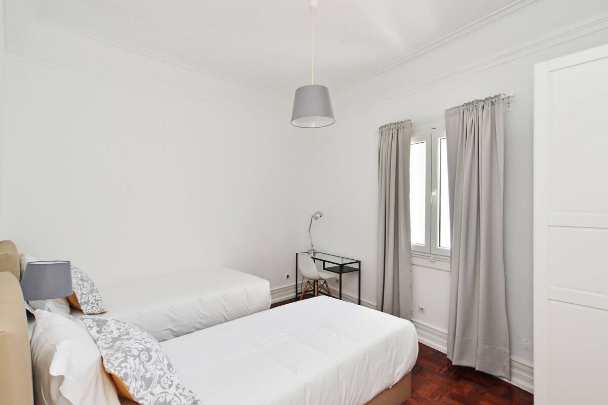 A bedroom with two beds, a desk with a chair, a hanging lamp, and a window with curtains.