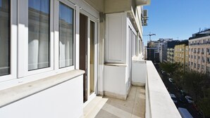 Een balkon met witte hekwerk, een raam met gordijn, en uitzicht op een stadsweg met gebouwen en geparkeerde auto's.