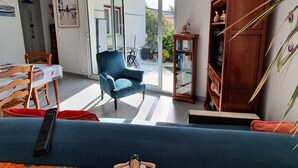A living room with a blue sofa, a teal armchair, a wooden bookshelf, and a dining area with a table and chairs.