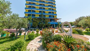 A multi-story hotel with balconies, surrounded by landscaped gardens and a large decorative sculpture. - Hotel Bahamas (Sarandë)