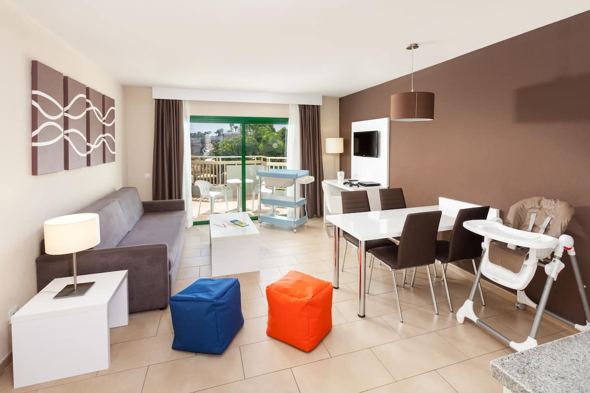 A modern hotel room with a balcony, a dining area, a sofa, a TV, and a baby changing station.