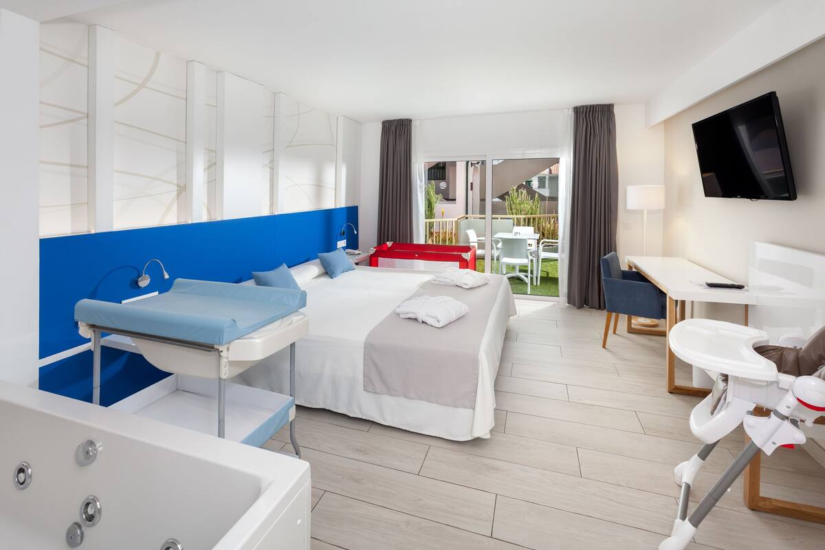 A modern hotel room with a large bed, a desk, a chair, a TV, a bathroom with a bathtub, and a balcony with outdoor seating.