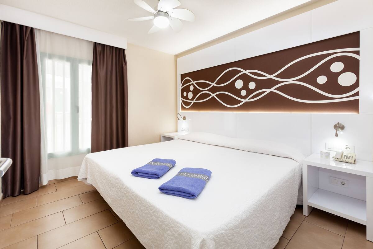 A hotel room with a large bed, two pillows, a bedside table with a lamp, a nightstand with a phone, and a decorative wall panel.