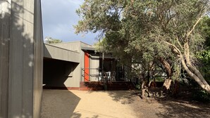 Un edificio dal design moderno, con facciata in cemento e una porta rossa. Si vede una scala che conduce a un'area coperta con posti a sedere all'aperto.