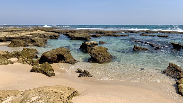 Una spiaggia rocciosa con acqua cristallina e un molo in lontananza.