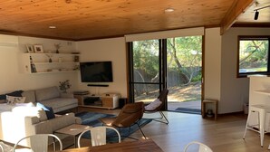 Un soggiorno moderno con soffitto in legno, una TV a schermo piatto, un divano, un tavolo da pranzo con frutta e vista sull'esterno.
