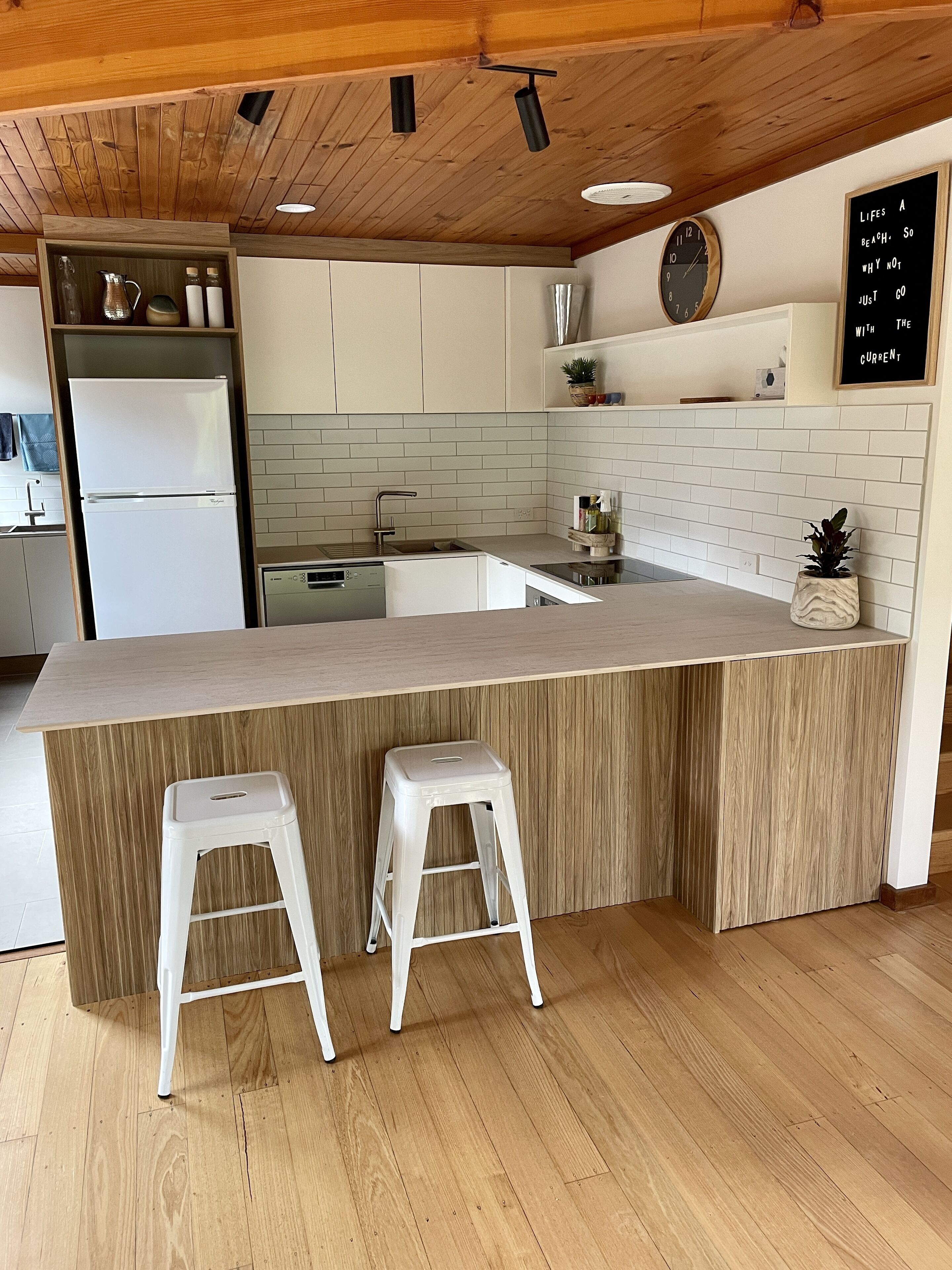 Uma cozinha moderna com teto de madeira, armários brancos, uma ilha grande com banquetas e um quadro-negro com uma frase motivacional.