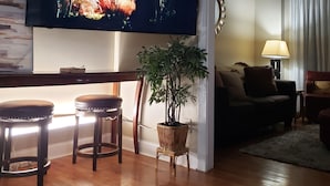 A living room with a flat-screen TV mounted on the wall, a wooden coffee table, two bar stools, a potted plant, a sofa, and a lamp. - Amazing W Marietta 5BR-3BA Relocation, Long Stay (Marietta)