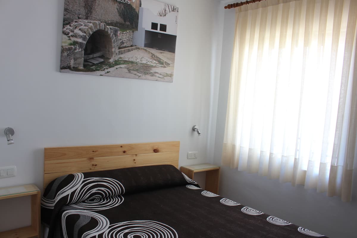 A bedroom with a wooden headboard, a bed with a patterned bedspread, a small bedside table, a nightstand, a wall-mounted light, and a window with sheer curtains.