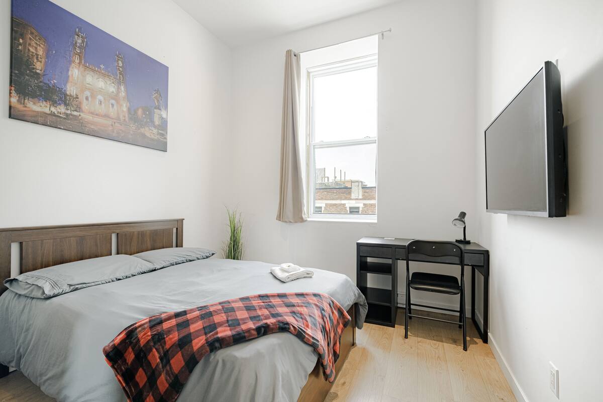 Un dormitorio con cama, escritorio, silla, televisor y ventana con vista a la ciudad.