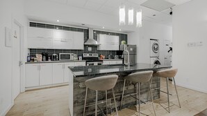 A modern kitchen with white cabinets, a marble island with bar stools, a built-in oven, and a washing machine in the adjacent room. - HoMa HomeStay Hotel by Olympic Stadium Montreal (Auberge/Hostel) (Montreal)