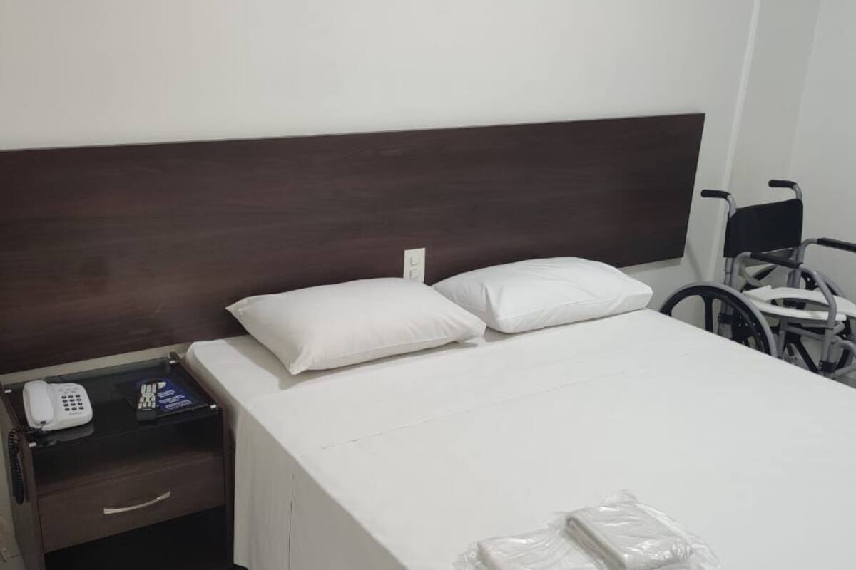 A hotel room with a single bed, a bedside table with a phone, and a wheelchair.