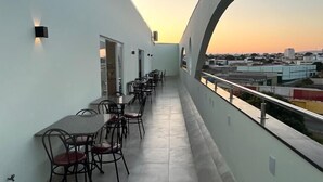 A balcony with a row of tables and chairs, a railing, and a view of the cityscape during sunset. - Hotel Rametta (Montes Claros)