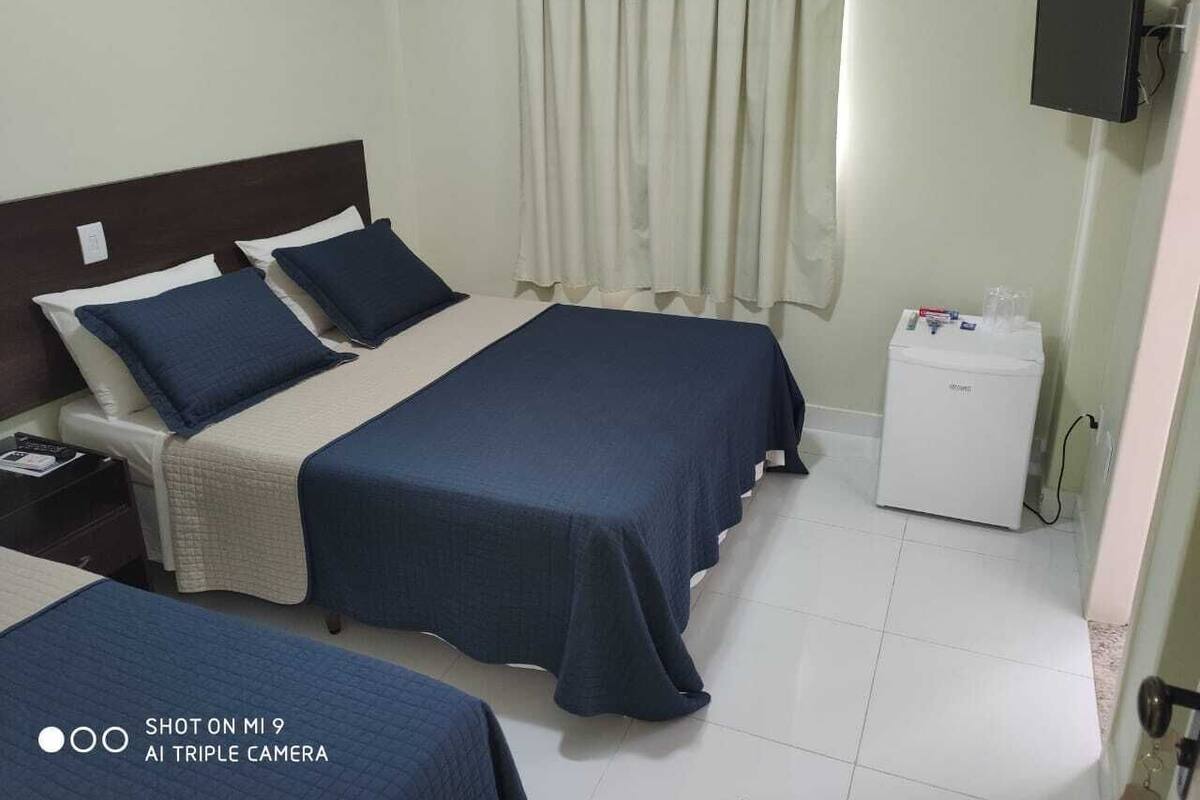 A hotel room with two beds, a small bedside table, a white bedside unit, and a curtain.