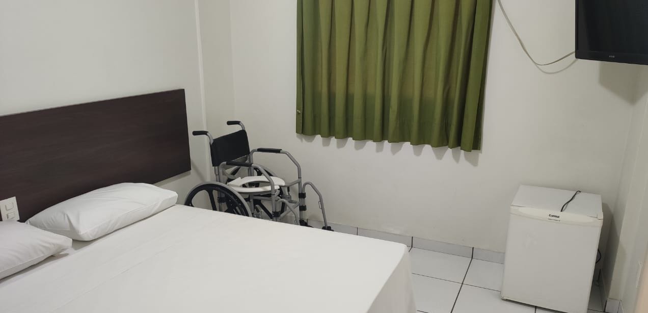 A hotel room with a bed, a wheelchair, a small white cabinet, and a green curtain.