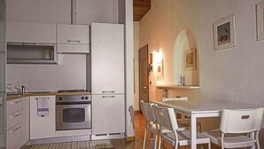 Een kleine keuken met een fornuis, oven en witte kasten. Er is een eettafel met stoelen en een zithoekje.