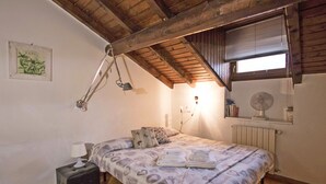 Een slaapkamer met een houten plafond, een bed met beddengoed met patroon, een nachtkastje met een lamp, een radiator en een raam met zonwering.