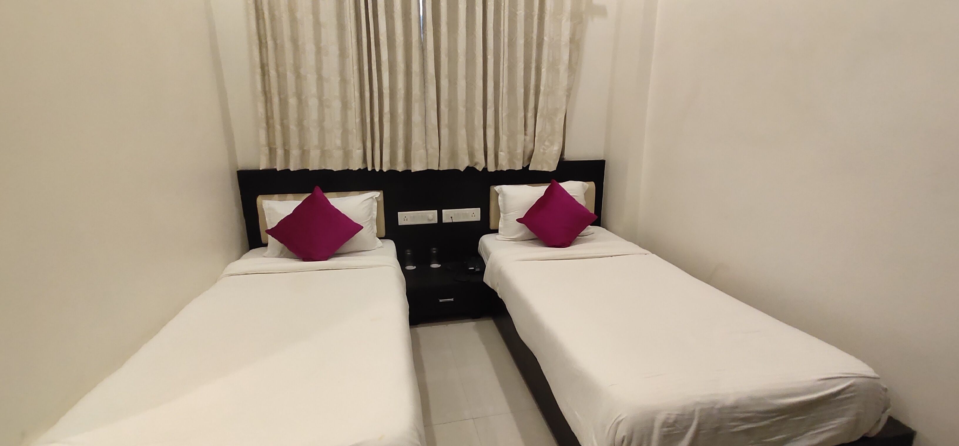 A small, clean room with two single beds, each with a white mattress and a purple pillow. There is a black headboard between the beds, and a window with curtains.