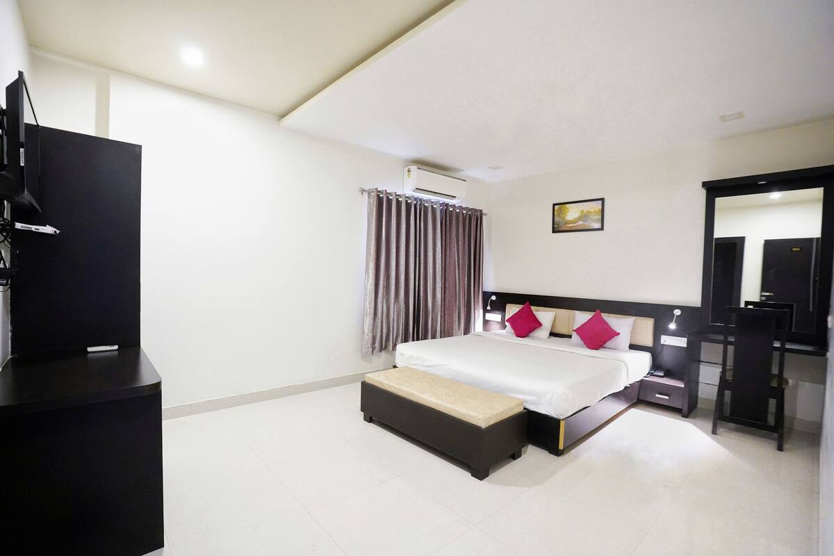 A hotel room with a bed, bedside table, a desk with a chair, an air conditioning unit, and a television.