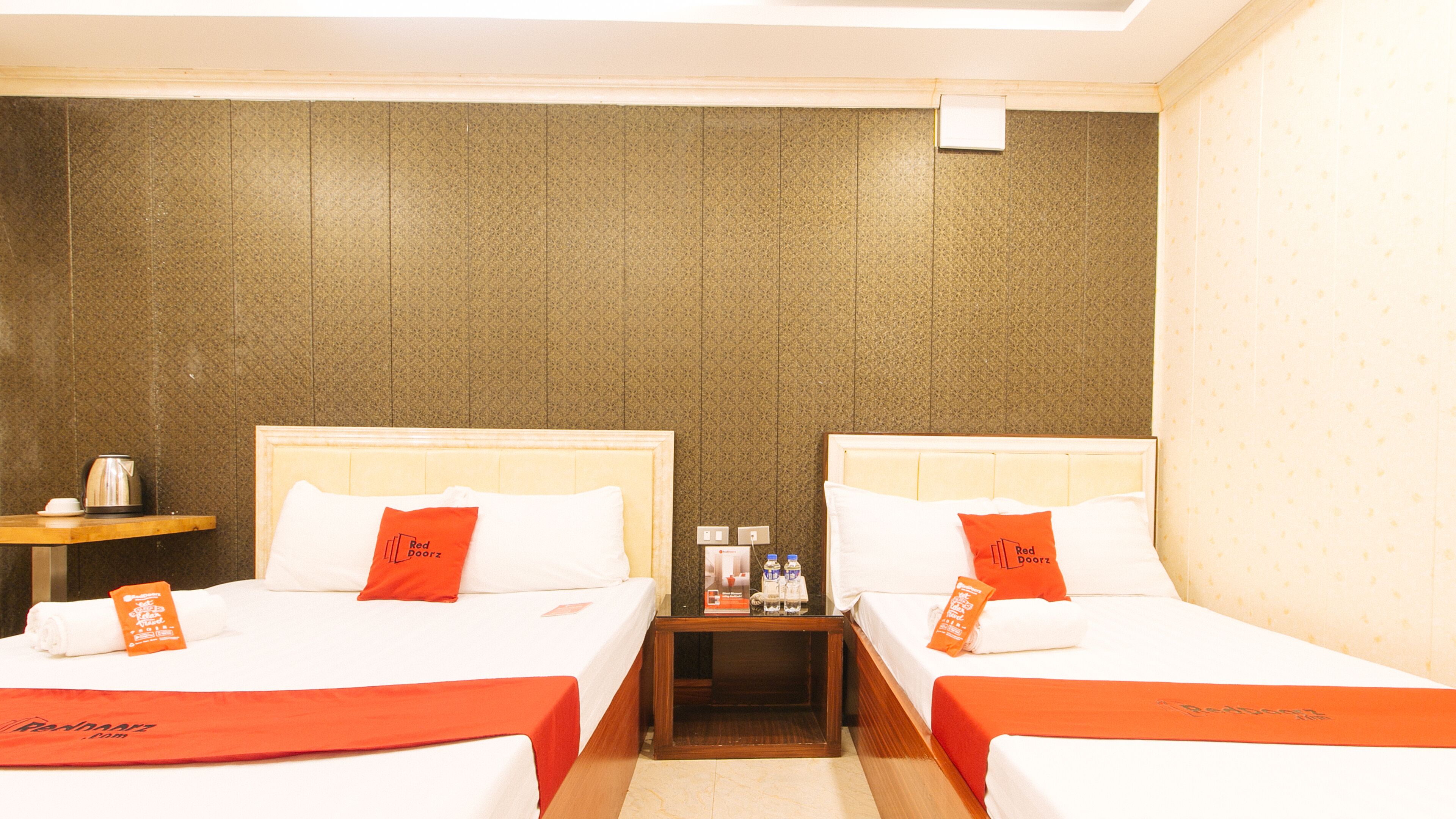 A hotel room with two beds, each with a red and white bedspread and matching pillows. There is a nightstand between the beds with a coffee pot and bottled water. The walls have a textured pattern, and there is a small table with a cup and a tissue box.