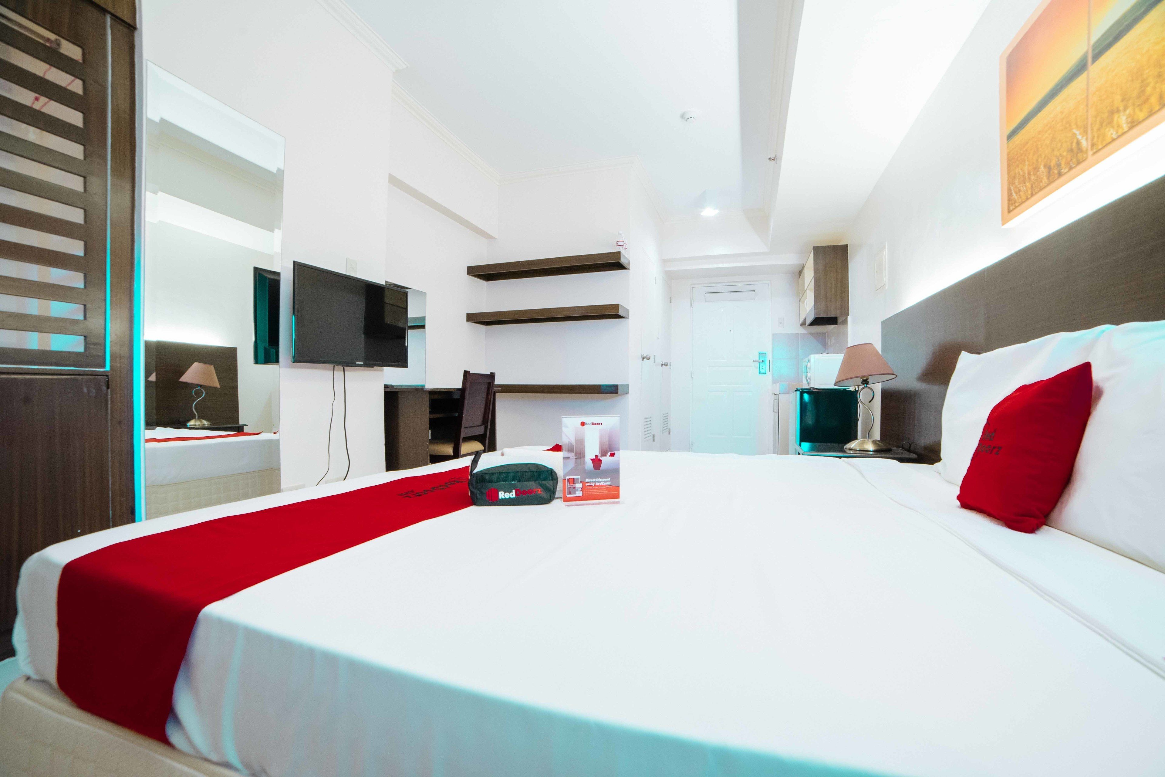 A modern hotel room with a large bed, a flat-screen TV, a desk with a lamp, and a bathroom visible in the background.