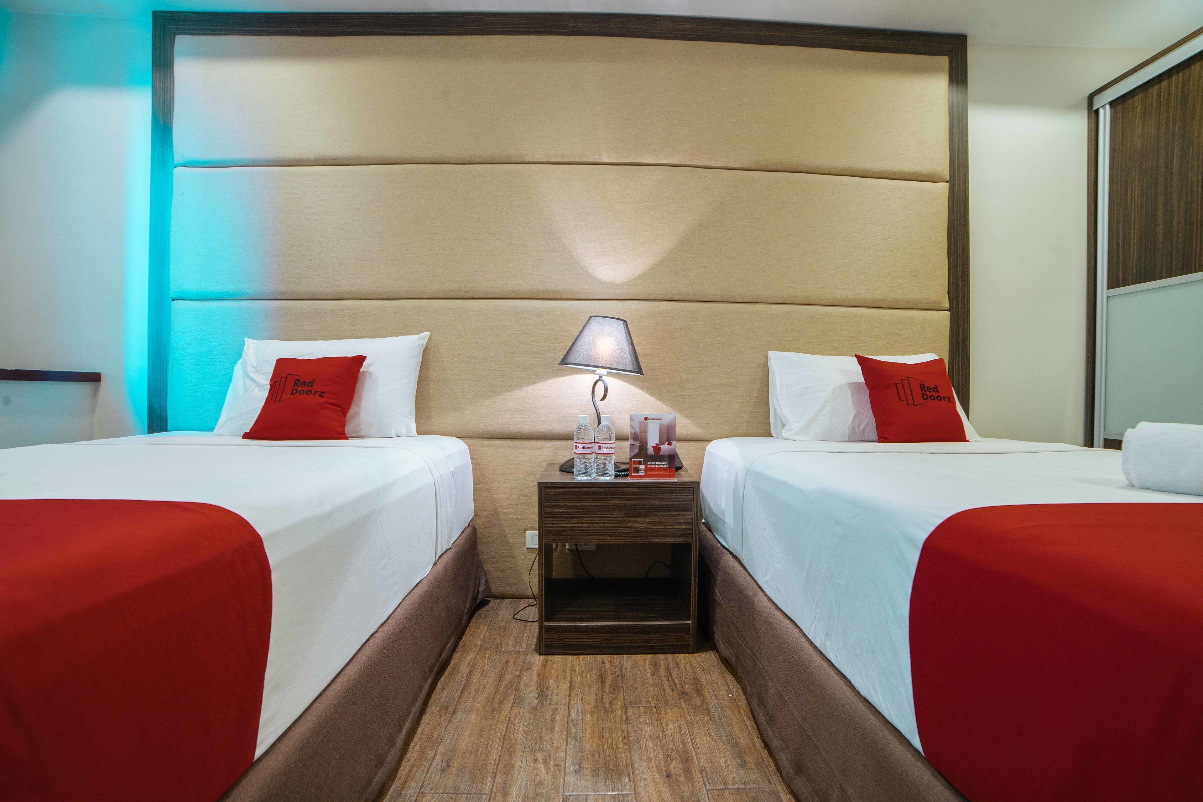 A hotel room with two beds, each with red bedding and pillows, a nightstand with a lamp, and a wooden headboard.