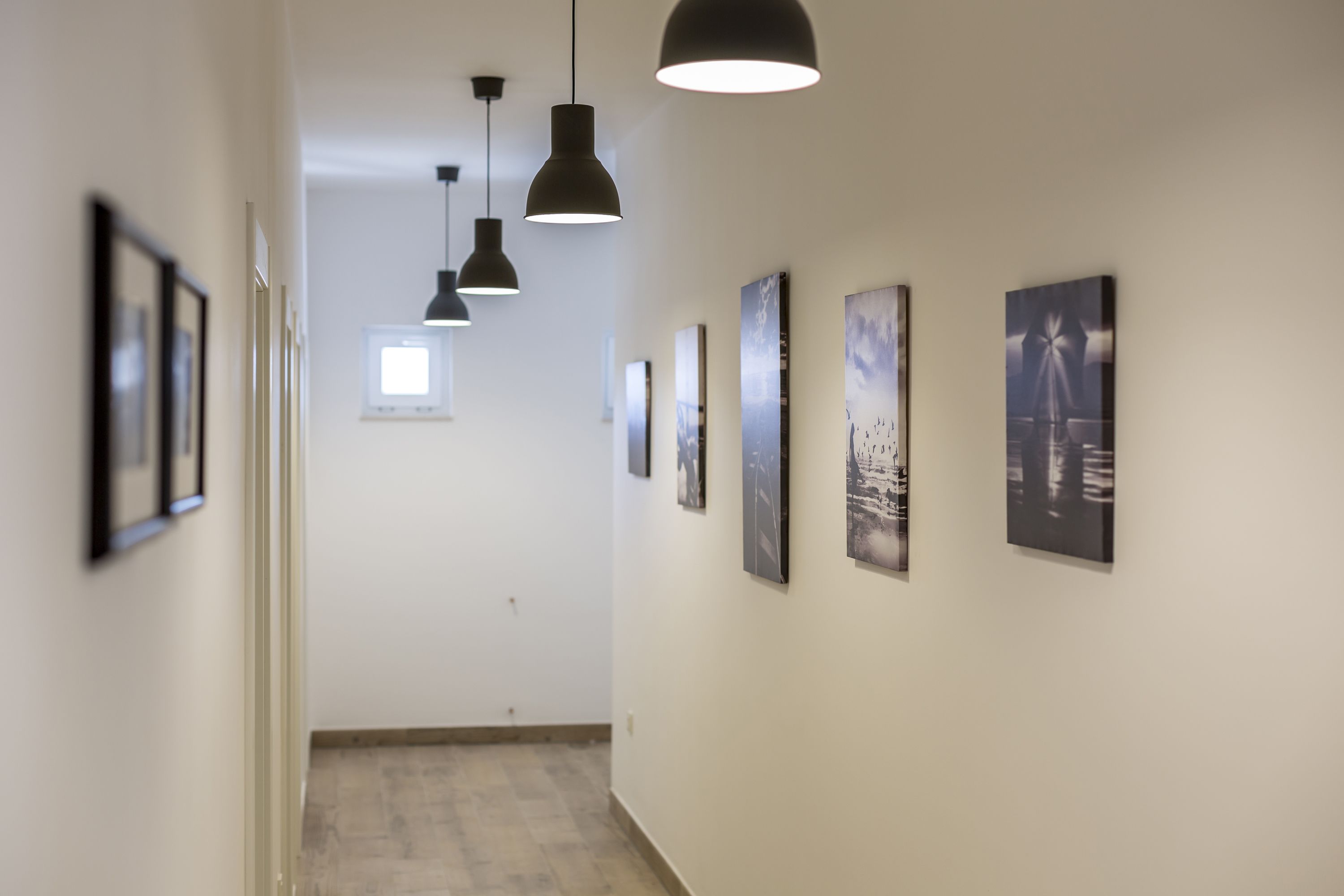 A hallway with framed pictures on the walls, pendant lights, and a window at the end.