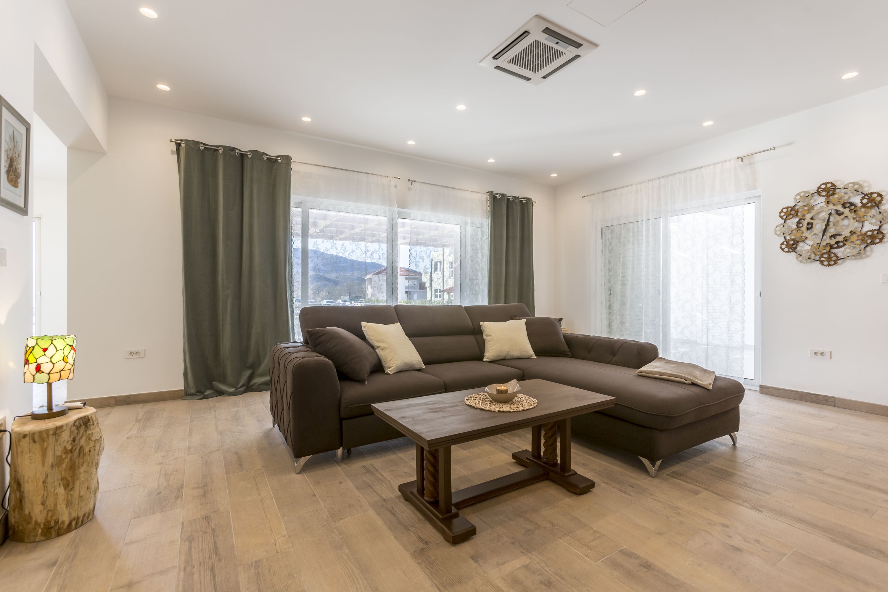 Un salon moderne avec un canapé d’angle, une table basse en bois et de grandes fenêtres avec des rideaux.