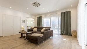 Una sala de estar moderna con un sofá, un reposapiés y una mesita. Se ve una ventana con cortinas y una puerta al fondo.