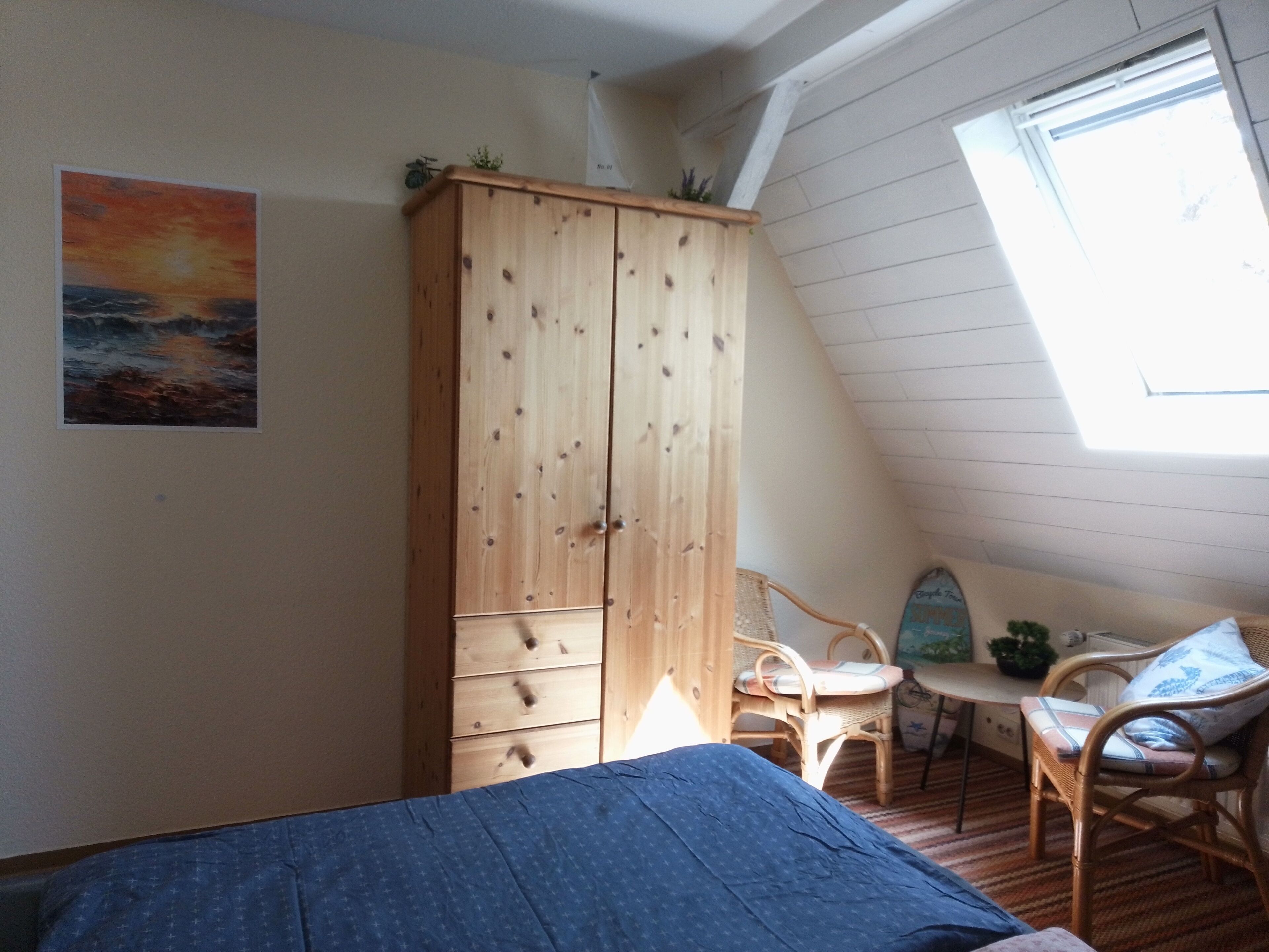 Ein Schlafzimmer mit Holzschrank, Bett mit blauer Decke, Korbsessel, Kleintisch und Fenster mit Blick zum Himmel.