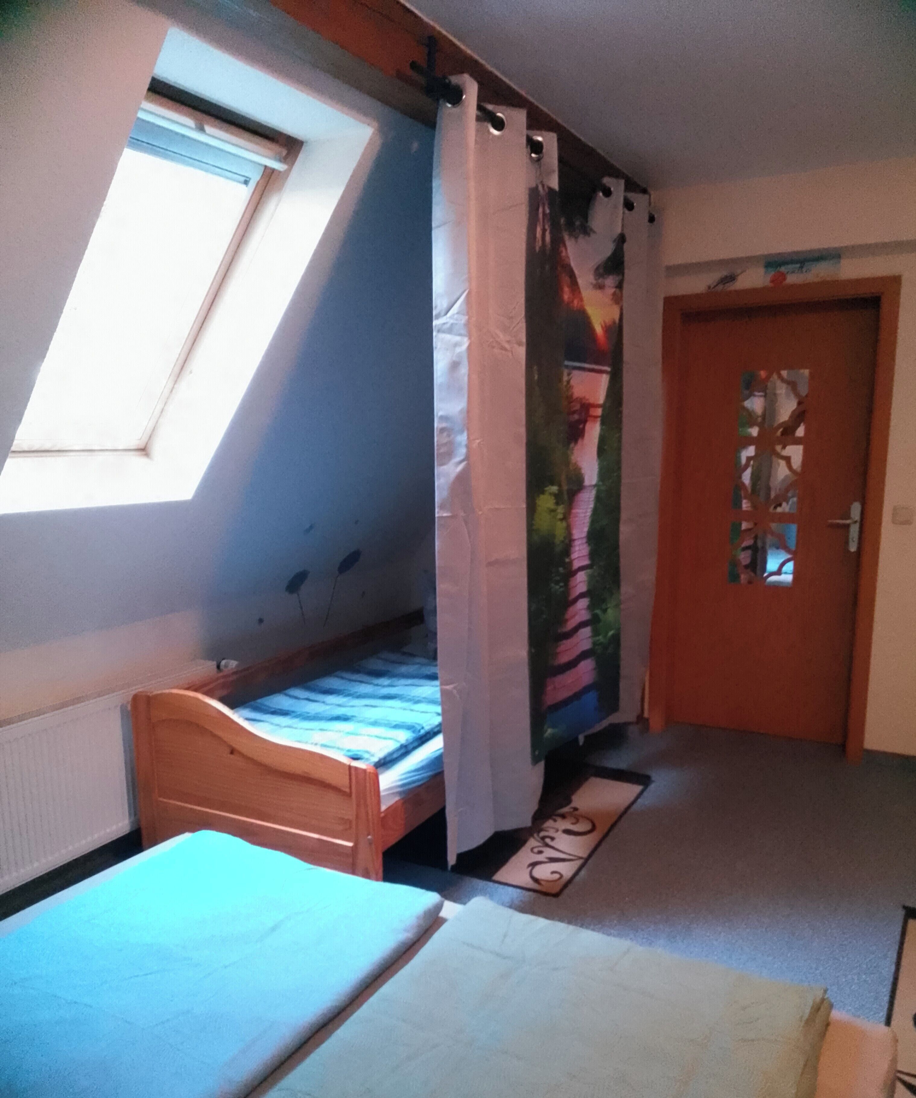 Ein Zimmer mit einem Einzelbett, einem Fenster mit Vorhang, einer Tür mit dekorativem Glasfeld und einem kleinen Teppich auf dem Boden.