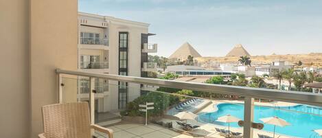 A balcony with wicker furniture, a view of a swimming pool, and pyramids in the background.