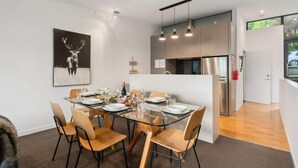 A modern dining area with a glass table, wooden chairs, and a framed deer artwork on the wall. - Happy Daze Central Queenstown Hot Tub (Queenstown)