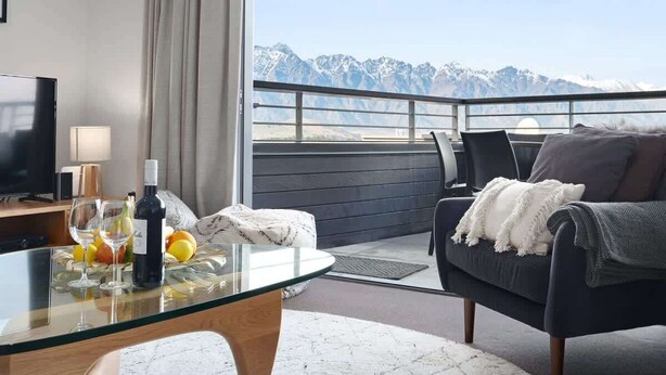 A modern room with a glass table, a sofa, and a view of snow-capped mountains. - Happy Daze Central Queenstown Hot Tub (Queenstown)