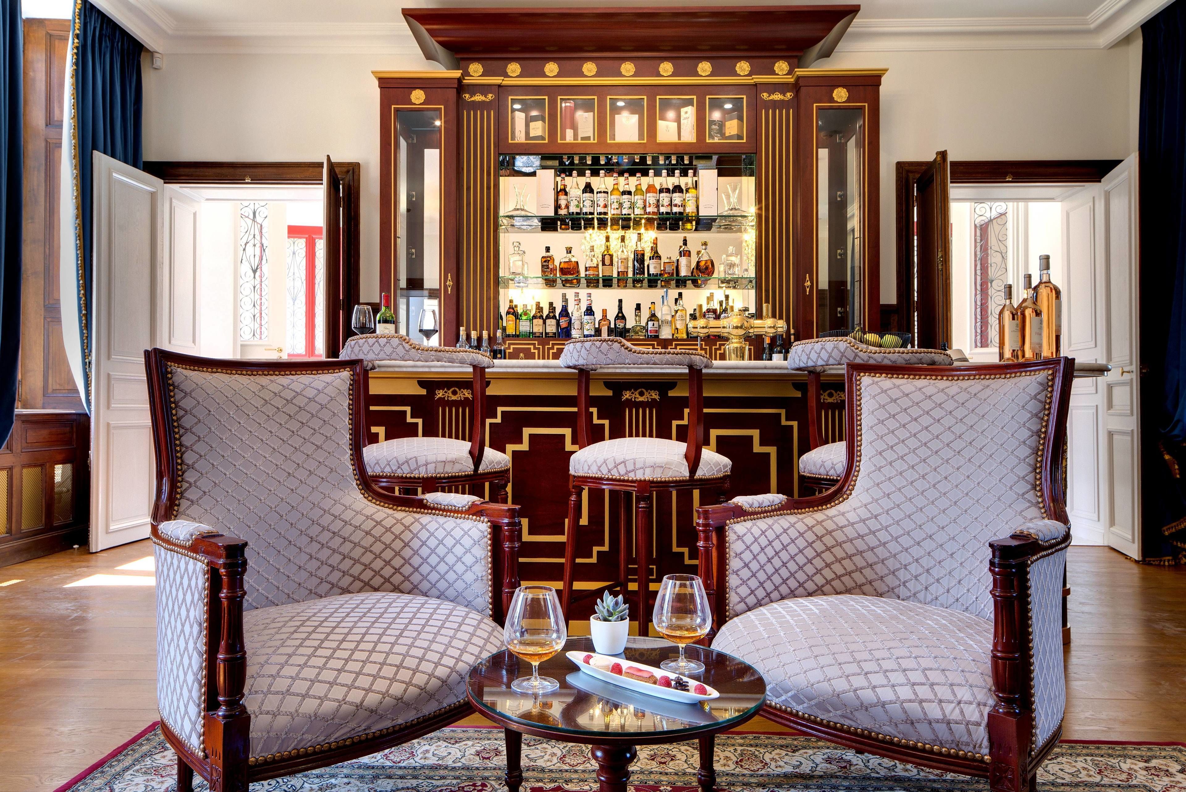 A bar area with a wooden cabinet displaying bottles, two upholstered chairs with cushions, and a round table with glasses and a plate.
