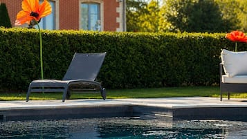 Una piscina con una silla de descanso y un banco, un seto y un edificio de ladrillo al fondo.