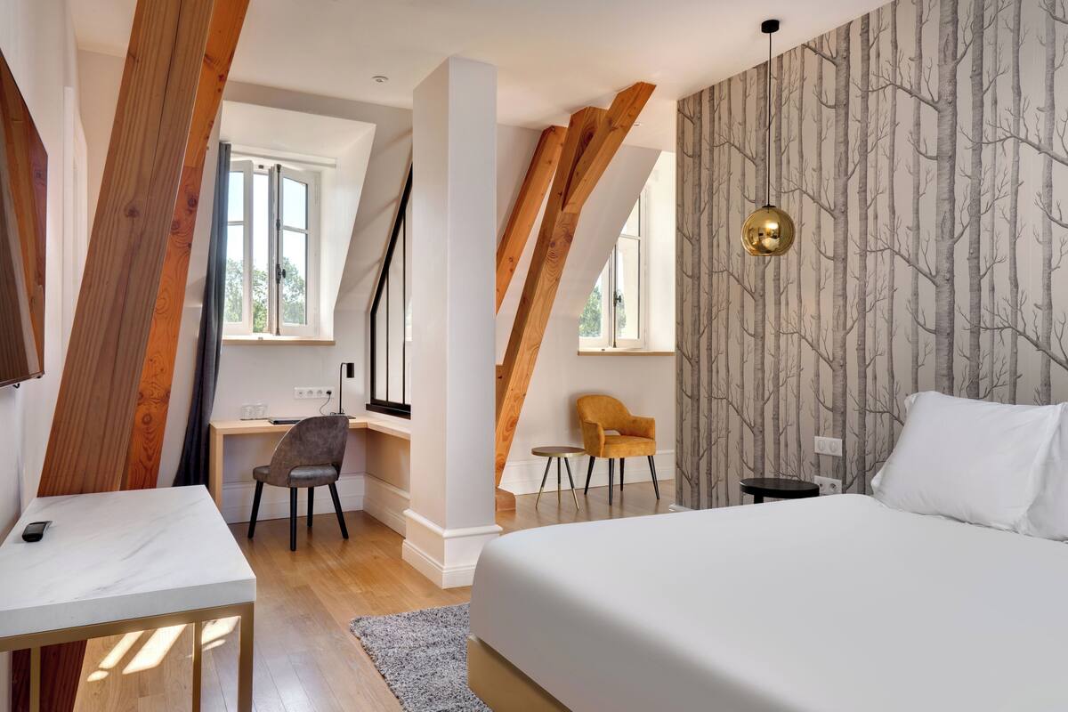 Une chambre avec un grand lit, un bureau et une chaise. Il y a une fenêtre donnant sur les arbres.