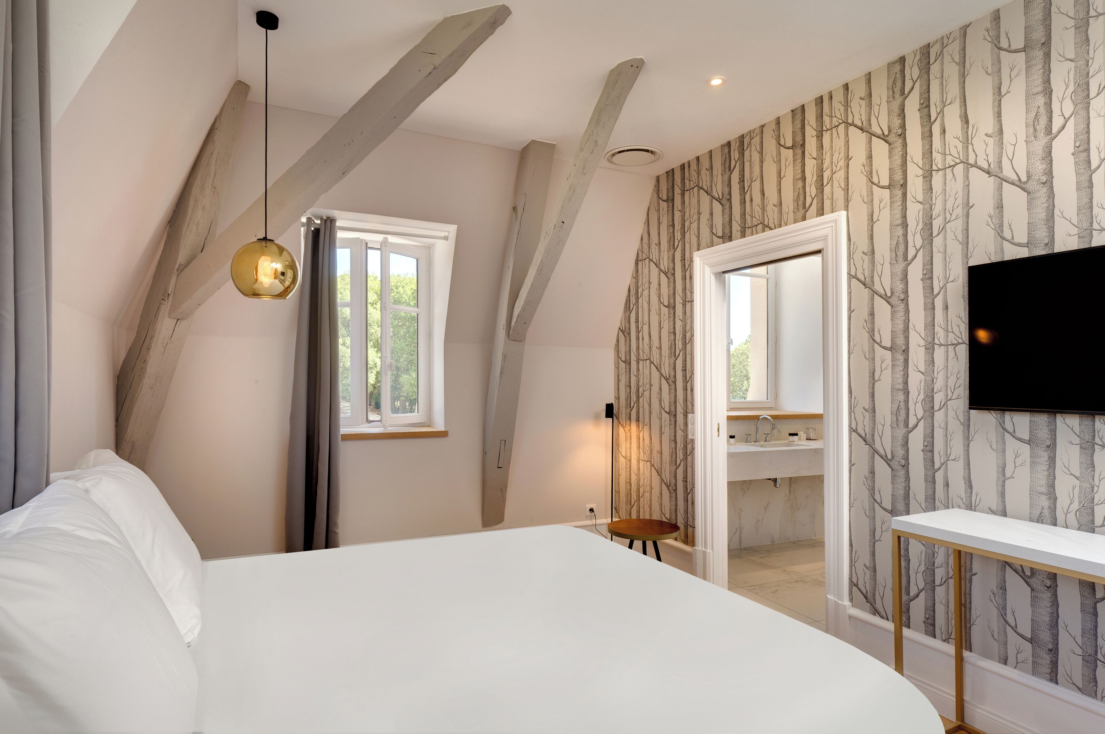 A bedroom with a sloped ceiling, a bed, a bedside table, a lamp, a TV, and a bathroom visible through an open door.