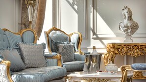 Une chambre luxueuse, avec un canapé bleu et doré richement décoré, une table blanche ornée d’un vase et de deux verres, et un buste sculpté posé sur un piédestal.