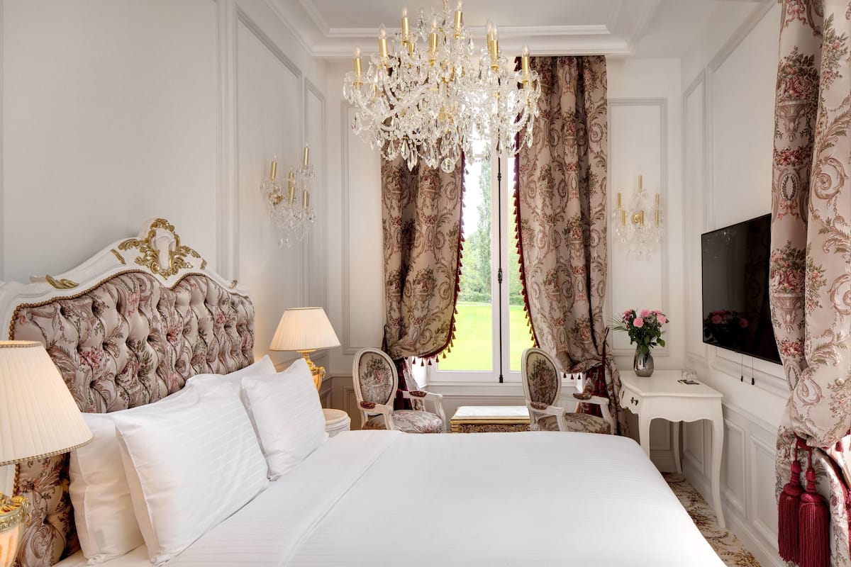 Une chambre à coucher luxueuse avec un grand lit, une tête de lit décorée, un lustre, des rideaux à motifs floraux, une télévision et une petite table sur laquelle est posé un vase rempli de fleurs.
