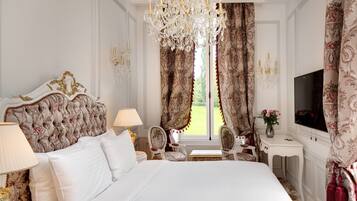 Een luxueuze slaapkamer met een groot bed, een versierd hoofdeinde, een kroonluchter, bloemen gordijnen, een televisie en een klein tafeltje met een bloemenvaas.