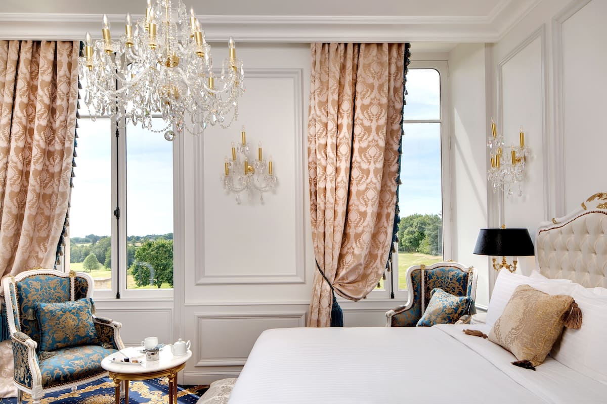 Une chambre à coucher luxueuse, dotée d’un grand lit, de fauteuils richement décorés, d’un lustre et offrant une vue sur l’extérieur.