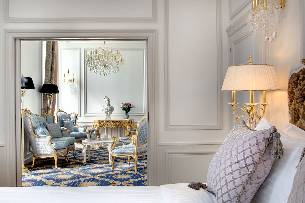 Une chambre à coucher luxueuse, dotée d’un grand lit, d’un coin salon avec des fauteuils élégants, d’un lustre et d’un décor classique aux couleurs blanc et or.