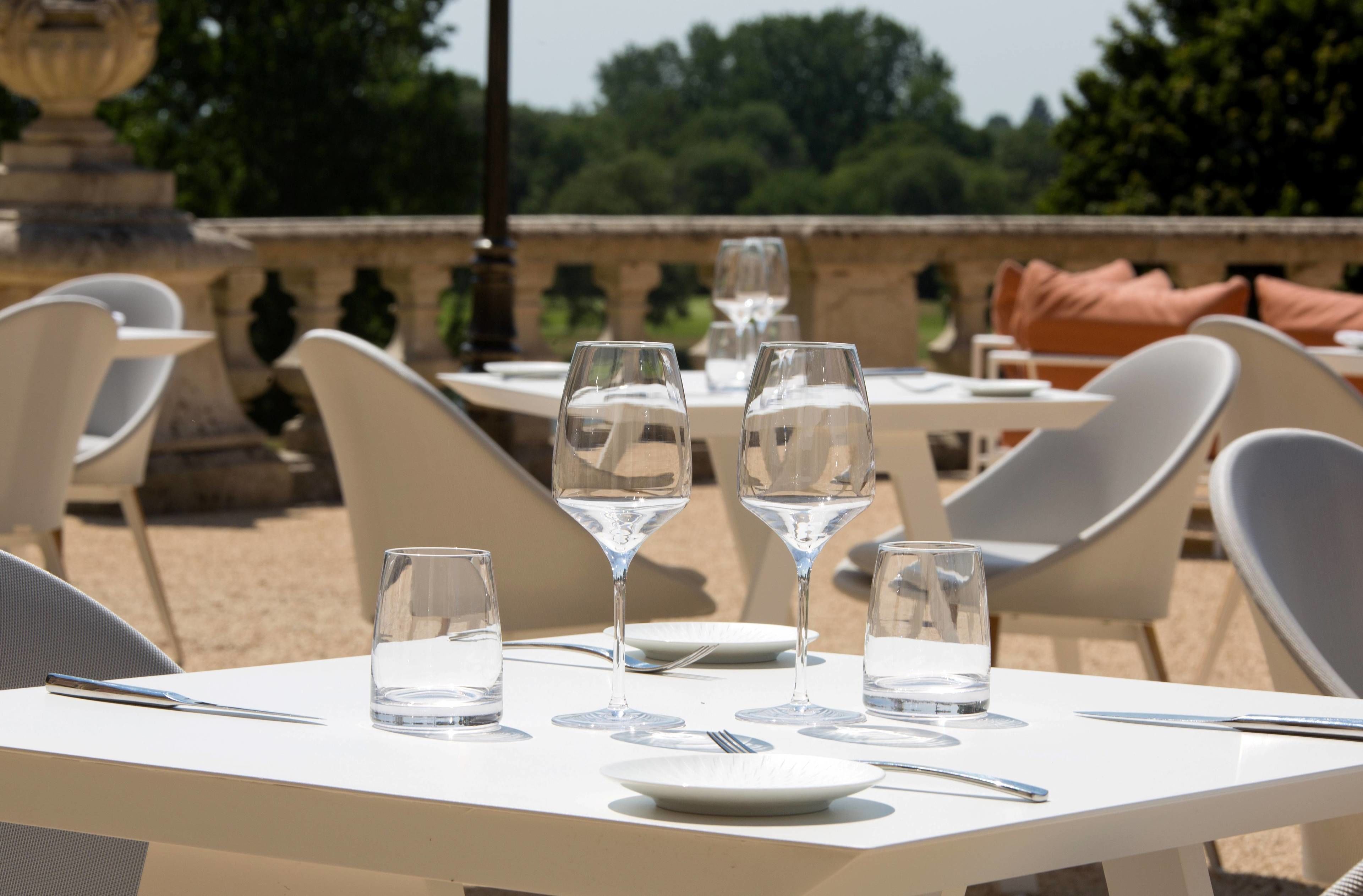 Une table dressée avec des verres à vin et des verres à eau, des assiettes blanches et des couverts, avec en toile de fond une vue sur la verdure et une balustrade en pierre.