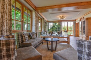 A spacious living room with a large sofa set, a round coffee table, and a view of greenery through large windows.