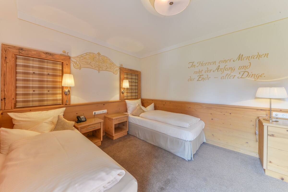 A hotel room with two beds, a wooden headboard, a bedside table with a lamp, a small desk, and a wall with a decorative quote.