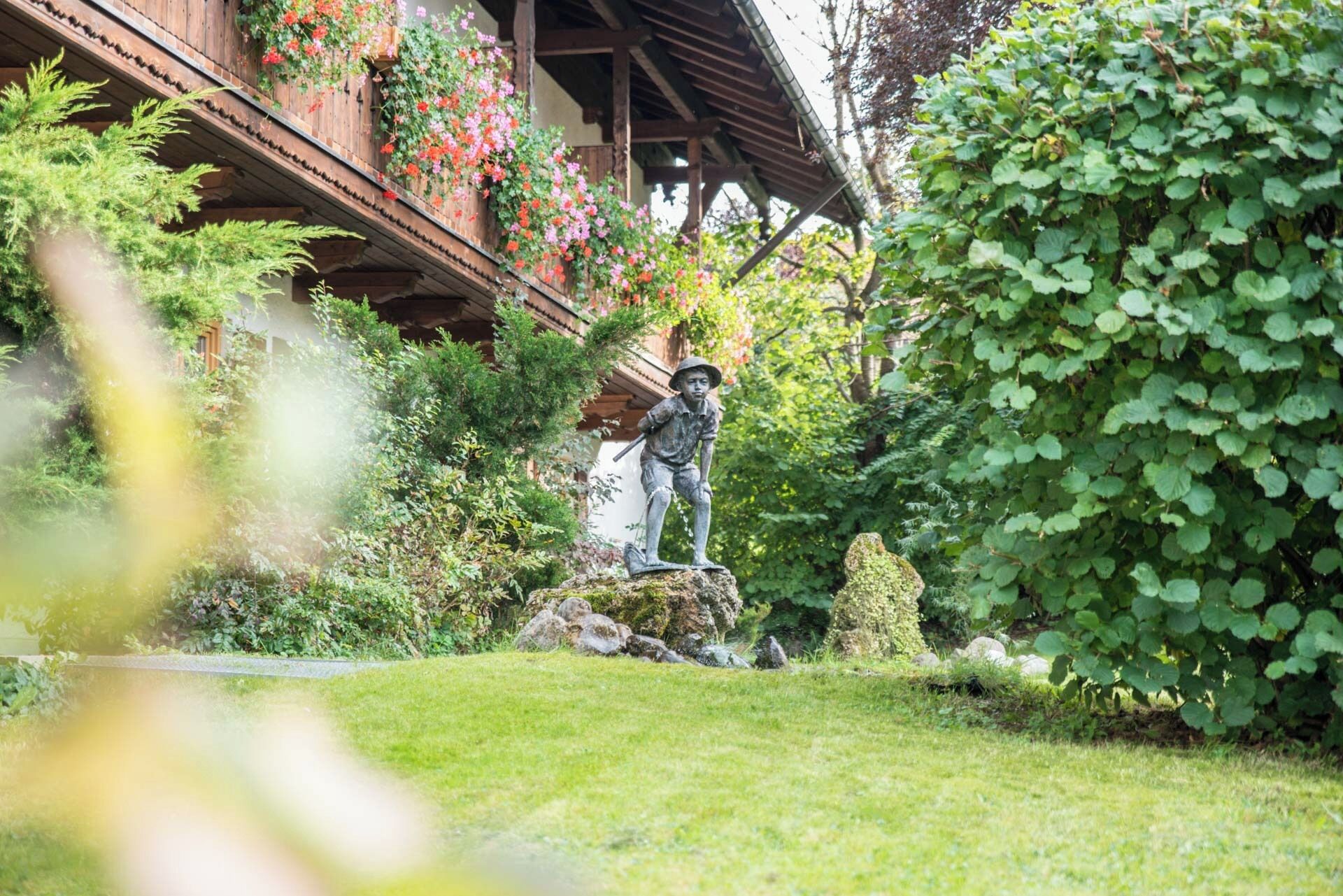Un jardín con una estatua de una persona, abundante vegetación y un edificio de madera con jardineras.