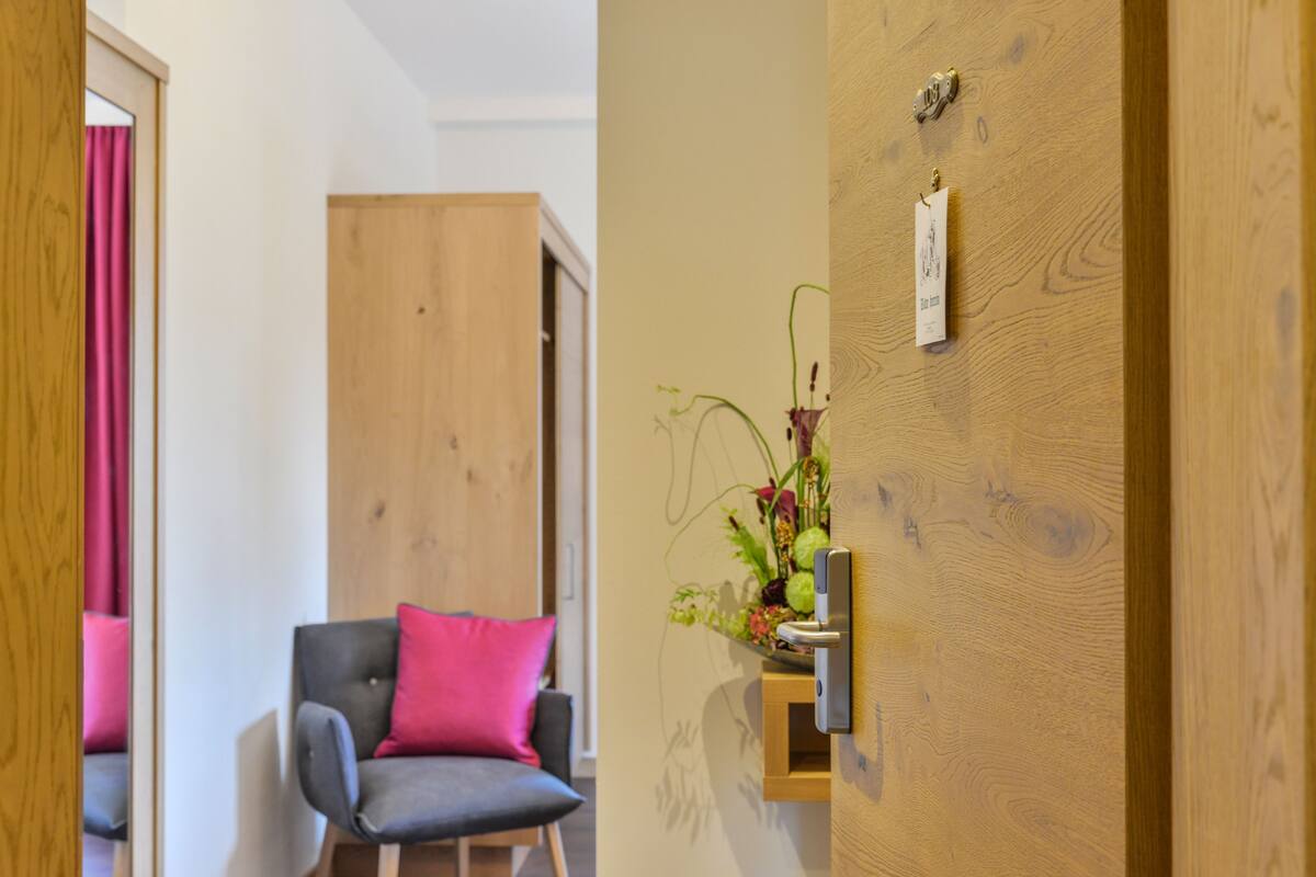A room with a wooden door, a grey chair with a pink pillow, and a small shelf with a plant.