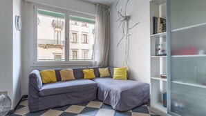 A room with a checkered floor, a grey sectional sofa with yellow cushions, a window with a view of buildings, a white bookshelf, and a wall-mounted light fixture. - Venezia - Lido Erizzo 15 (Venice)